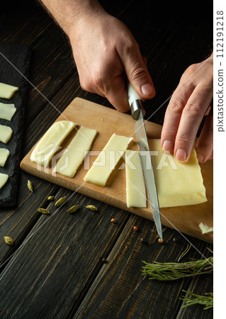 The cook hands with a knife cut Mozzarella cheese on a cutting board in a restaurant kitchen for preparing snack sandwiches. Delicious snack for lunch 112191218