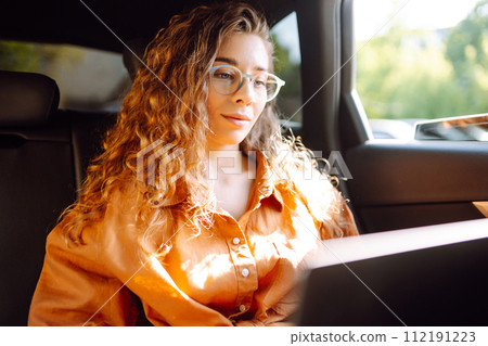 A young curly woman with a laptop sitting in back seat of a car. Beautiful woman uses laptop in car. 112191223