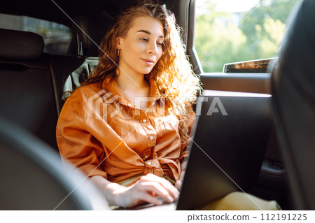 A young curly woman with a laptop sitting in back seat of a car. Beautiful woman uses laptop in car. A young curly woman with a laptop sitting in back seat of a car. Beautiful woman uses laptop in car. 112191225