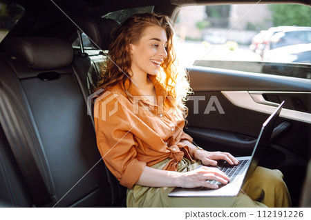 A young curly woman with a laptop sitting in back seat of a car. Beautiful woman uses laptop in car. 112191226