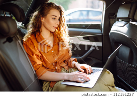 A young curly woman with a laptop sitting in back seat of a car. Beautiful woman uses laptop in car. A young curly woman with a laptop sitting in back seat of a car. Beautiful woman uses laptop in car. 112191227