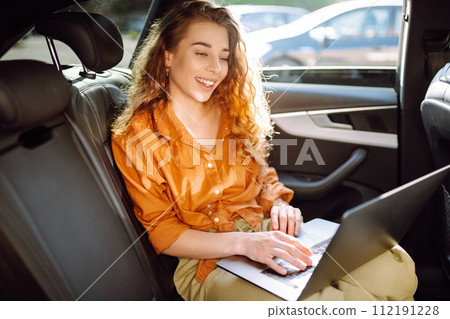 A young curly woman with a laptop sitting in back seat of a car. Beautiful woman uses laptop in car. 112191228