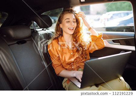 A young curly woman with a laptop sitting in back seat of a car. Beautiful woman uses laptop in car. 112191229