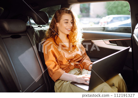 A young curly woman with a laptop sitting in back seat of a car. Beautiful woman uses laptop in car. A young curly woman with a laptop sitting in back seat of a car. Beautiful woman uses laptop in car. 112191327