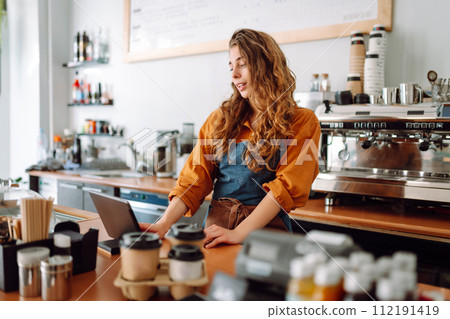 Young woman in apron with digital tablet at bar counter of coffee shop takes order. Small business. 112191419