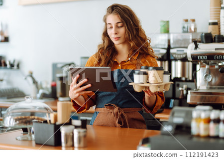 Handsome manager of coffee shop with curly hair tablet in hands gives takeaway coffee in coffee shop Handsome manager of coffee shop with curly hair tablet in hands gives takeaway coffee in coffee shop 112191423