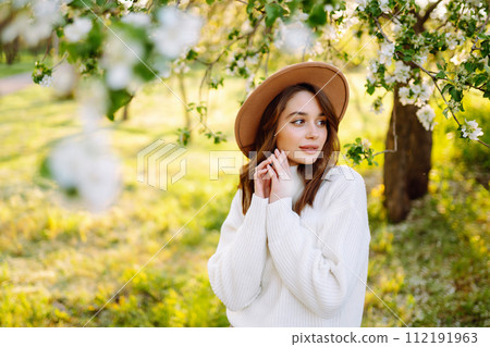Portrait of Woman In hat posing near flowering tree. Smiling young woman enjoying smell of flowers. 112191963