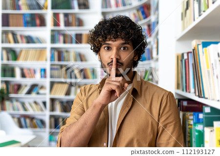 A young adult male with curly hair placing a finger on his lips, signaling quietness amidst a backdrop of bookshelves filled with various books. He portrays a sense of silence and focus, typical of a 112193127