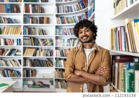 A cheerful young man with curly hair stands confidently with his arms crossed in a well-stocked library. He projects a vibe of intelligence and positivity amidst a collection of books. A cheerful young man with curly hair stands confidently with his arms crossed in a well-stocked library. He projects a vibe of intelligence and positivity amidst a collection of books. 112193138