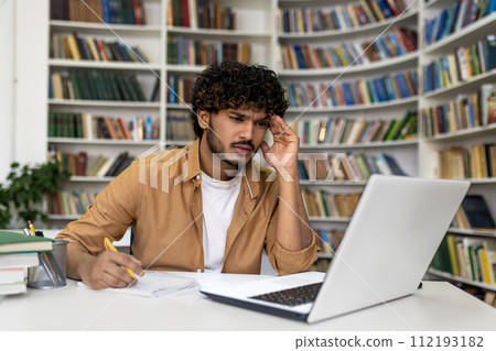 A focused young man is immersed in his studies at the library, surrounded by books, with a laptop for assistance. He seems to be preparing for exams, reflecting the life of a university or academy 112193182