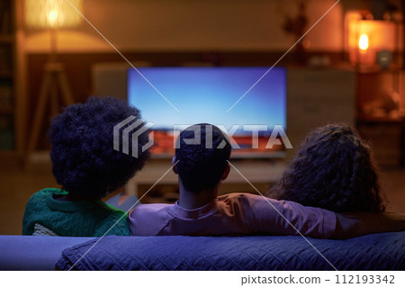 Back view of three people sitting on couch together and watching TV in dark room Back view of three people sitting on couch together and watching TV in dark room 112193342