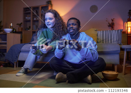 Full length portrait of Black man and Caucasian woman playing video games in living room sitting on floor and holding controllers 112193346