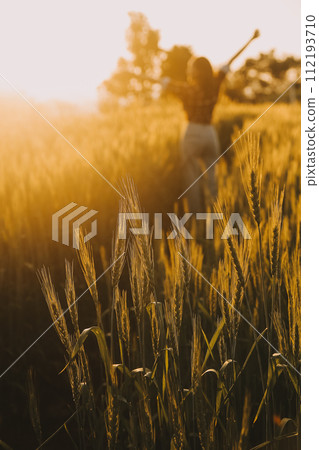 Young pretty woman in red summer dress and straw hat walking on yellow farm field with ripe golden wheat enjoying warm evening. Young pretty woman in red summer dress and straw hat walking on yellow farm field with ripe golden wheat enjoying warm evening. 112193710