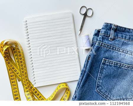 Tailoring and fashion design concept with denim jeans, yellow measuring tape, scissors, thread, and notepad with blank page for text on white background for planning and alterations. Top view. 112194568