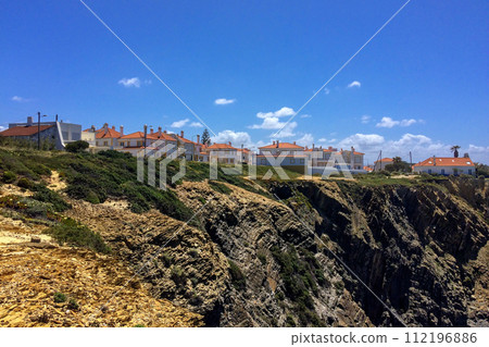 Coastal town with orange-roofed houses on a cliff, rocky terrain in the foreground, and a clear blue sky. 112196886