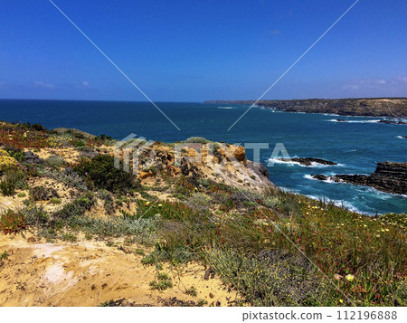 A coastal landscape with rocky cliffs, greenery, and a clear blue sky. The ocean waves crash against the rocks. A coastal landscape with rocky cliffs, greenery, and a clear blue sky. The ocean waves crash against the rocks. 112196888