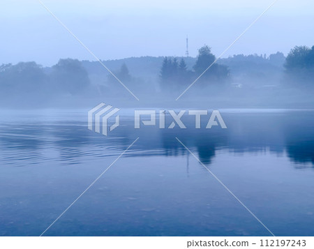 Morning fog on the lake in the early misty morning. The trees are reflected in the water. 112197243