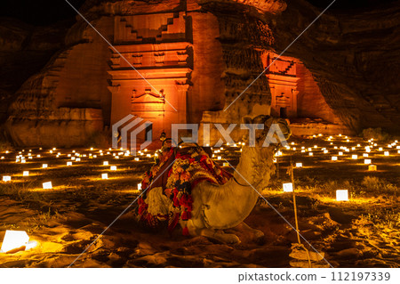 Camel laying in front of ancient tombs of Hegra city illuminated during the night, Al Ula, Saudi Arabia 112197339