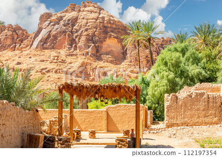 Al Ula ruined old town streets with straw sunshade tent and palms with mountains in the background, Saudi Arabia 112197354