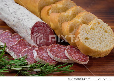 Spanish Fuet sausage salami with rosemary and french baguette on a cutting board. 112197409