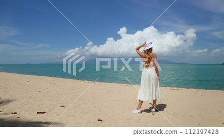 Young woman stands on sandy beach looking on ocean. Female tourist in white dress on summer vacation. Lifestyle holiday. Clear blue sky above calm sea in background, tranquil tropical seaside scene. Young woman stands on sandy beach looking on ocean. Female tourist in white dress on summer vacation. Lifestyle holiday. Clear blue sky above calm sea in background, tranquil tropical seaside scene. 112197428