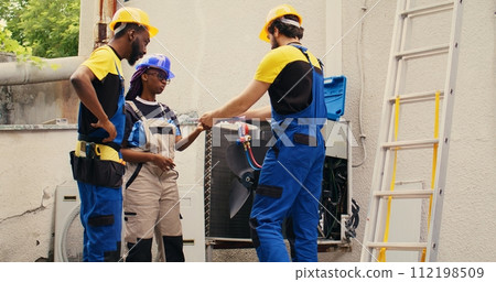 Diverse team of experienced mechanics brainstorming possible ways to recondition defective outdoor condenser. Skillful repairmen commissioned by client to patch up broken air conditioner 112198509