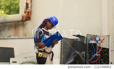 Proficient expert servicing outdoor air conditioner internal components, checking refrigerant level and calibrating thermostats. Capable specialist looking over maintenance plan on clipboard Proficient expert servicing outdoor air conditioner internal components, checking refrigerant level and calibrating thermostats. Capable specialist looking over maintenance plan on clipboard 112199063