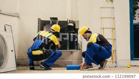 Teamworking competent mechanic opening up rusty out of service HVAC system to replace it with new performant outside condenser after draining refrigerant and replacing ductwork 112199097
