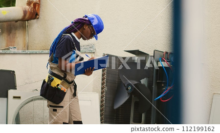 Capable electrician servicing external air conditioner internal components, checking freon level and calibrating thermostats. African american mechanic looking over maintenance plan on clipboard Capable electrician servicing external air conditioner internal components, checking freon level and calibrating thermostats. African american mechanic looking over maintenance plan on clipboard 112199162