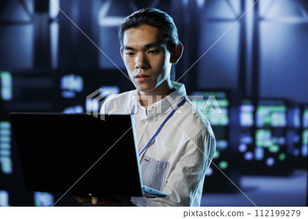 Qualified engineer between server hub rows providing processing resources for businesses worldwide. Certified expert fixing data center mainframes tasked with managing massive databases 112199279
