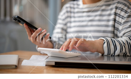 A cropped shot of a woman using her smartphone and a calculator at a desk, calculating her bills. 112201334