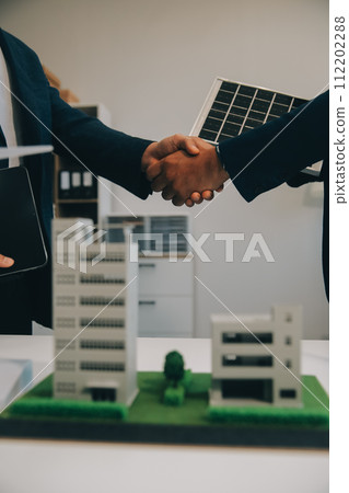 Two Young Engineers Expertise in Solar Cell Installation Meetings and Discussion in The Job. Planning to Install Solar Photovoltaic Panels on Roof Top in The Office Room with Factory Building Plan. 112202288