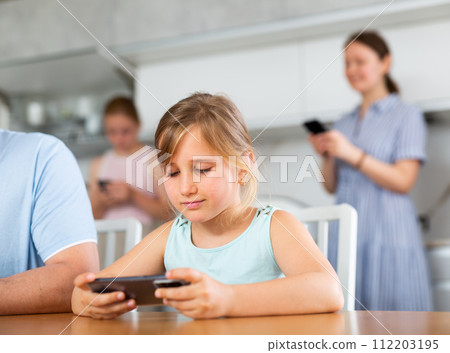 Preschooler girl sitting near kitchen table with phone in hands and playing online game Preschooler girl sitting near kitchen table with phone in hands and playing online game 112203195