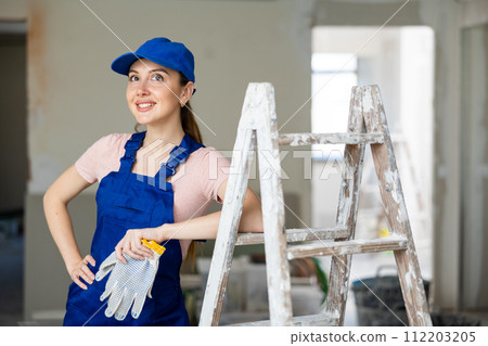 Portrait of positive builder woman in blue overalls next to stepladder 112203205