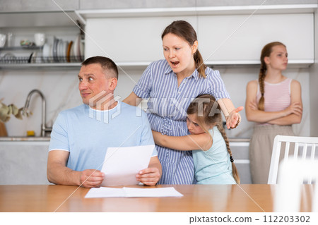 Angry woman chiding upset man sitting with papers in home kitchen 112203302