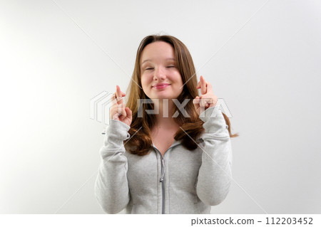 Intrigued woman in t-shirt praying with crossed fingers and looking away over grey background 112203452