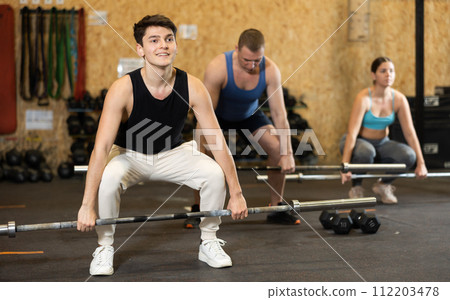 Energetic young male athlete practicing sit-ups with rod near other people gym 112203478