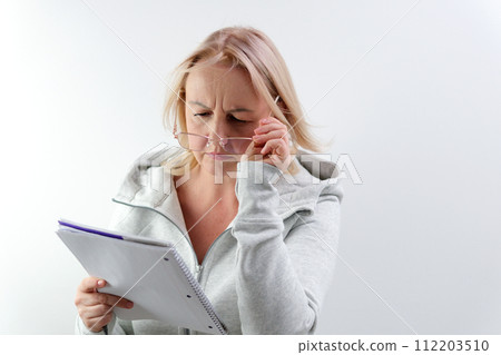 Middle-aged woman blonde writes on notebook with pen glasses in gold frames in studio checking eyesight falling links due to age self-care prescribing medications coming up with ideas 112203510