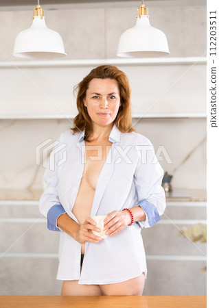 Topless woman in underpants and shirt stands in kitchen and covers bare chest with cloth of jacket. 112203511