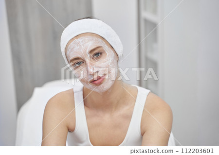 Young woman with white clay mask enjoys spa treatment in luxurious salon 112203601