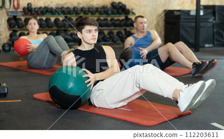Strong young man performing exercise for abs core with medicine ball on yoga mat during group workout in CrossFit gym center 112203611