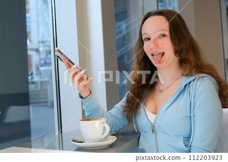 stick out tongue young teenage girl sits by the window with coffee and phone in hands looks into the frame showing tongue funny photos interesting emotion adolescence young woman 112203923