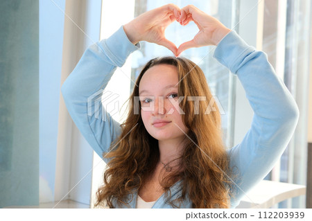 tender european girl makes heart with hands smiles sits at table by the window smile joy happiness convey love show heart on right at left and at top space for text advertising blue sports blouse 112203939