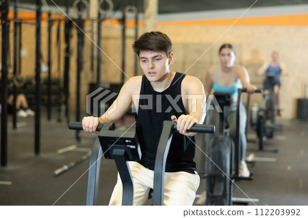 Dedicated active strong young man doing cardio workout on stationary bicycle as hard cross-training in gym indoors 112203992