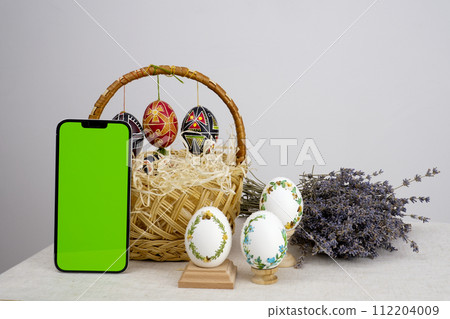 easter composition with phone with white chroma key screen for advertising basket of eggs with embroidery eggs painted lavender flowers wooden coasters on a white background Easter holiday easter composition with phone with white chroma key screen for advertising basket of eggs with embroidery eggs painted lavender flowers wooden coasters on a white background Easter holiday 112204009