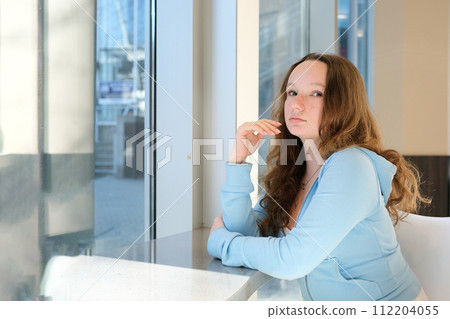 Depressed, sad woman at home looking out window for mental health, depression or heartbreak. Young gen z girl alone in apartment with lonely or bored face and thinking of problem or future life 112204055