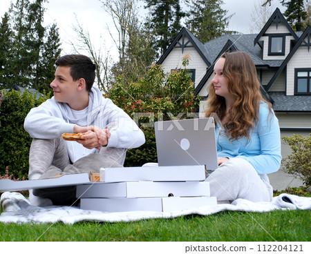A young girl and a boy look away with a slice of pizza and a laptop in their hands on the street, satisfied they waited for the food to be delivered. Very tasty Good time with friends 112204121