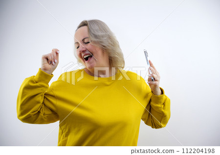 middle-aged adult woman dancing in bright clothes in the studio in the hands of a phone with a white screen space for text advertising positive emotions 112204198