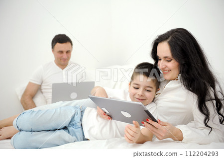 latest technology gadgets mom and son playing on tablet father sitting on white bed working on laptop remote work freelancer vacation family combine work and spend time with family together 112204293