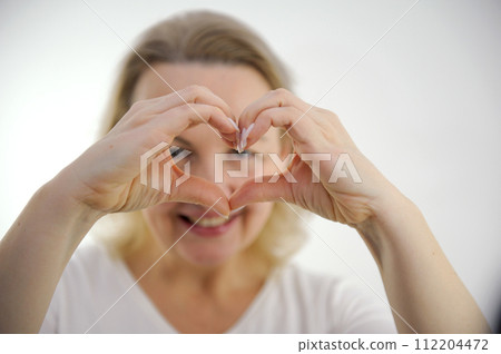 a woman shows her heart with her hands a cheerful look through her hands at the camera white background interest fun joy pleasant emotions happiness 112204472